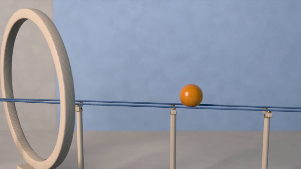 La bola rueda por dos cuerdas tensadas sobre postes de madera; detrás queda un aro grande de haya y una pared azul.